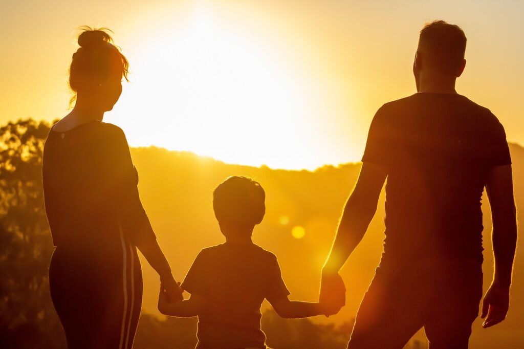 「家庭環境」をイメージした、両親と子供が手をつなぎ夕日を眺めている後ろ姿